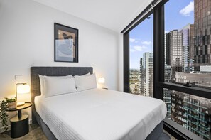 Ein modernes Hotelzimmer mit einem großen Bett, zwei Nachttischen mit Lampen, einem gerahmten Kunstwerk über dem Bett und Blick auf die Stadt durch große Fenster.