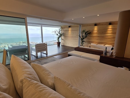 A modern hotel room with a large bed, a seating area with a view, a small table with a bottle and glass, and a wooden wall.. ‘Sitges Suites - Premium’ Apartment with Sea Views, Wi-Fi, and Air Conditioning
