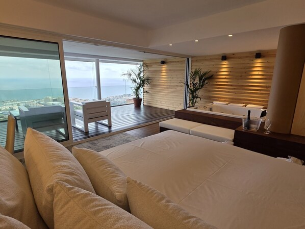 Una habitación de hotel moderna con una cama grande, un área de descanso con vistas, una mesita con una botella y un vaso, y una pared de madera.