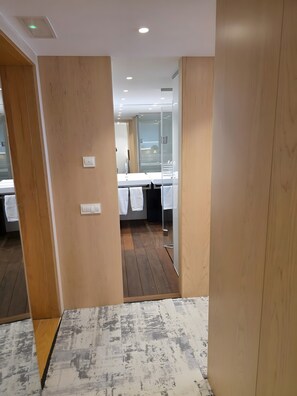 Habitación de hotel con suelo de madera, pared blanca, baño con mampara de vidrio y cama con ropa blanca.