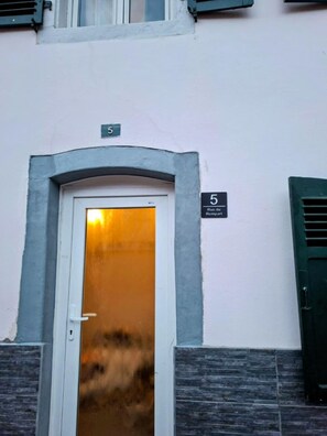 Entrada d'un edifici amb una porta blanca, un senyal amb el número 5 a dalt i una petita finestra amb persianes.