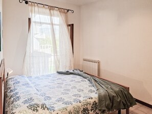 Une chambre à coucher avec un lit, un radiateur et une fenêtre ornée de rideaux légers.
