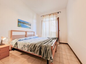 Un dormitori amb un llit, una tauleta de nit, una làmpada, un quadre a la paret i una finestra amb cortines.