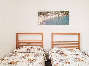 Een kamer met twee bedden, elk met een houten hoofdbord en passende kussens, en een schilderij van een uitzicht op zee aan de muur.