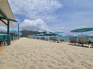 Una playa con filas de tumbonas y sombrillas, arena dorada y una vista al mar y montañas al fondo.