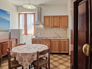 Una cocina compacta con armarios de madera, un salpicadero de azulejos, una mesa de comedor con mantel con motivos coralinos y una ventana con cortinas.