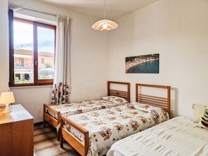 Un dormitorio con dos camas, un escritorio, una lámpara, una ventana con cortinas y una pintura con vista al mar.