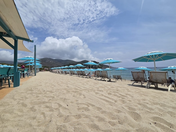 Una spiaggia con file di lettini e ombrelloni, una spiaggia sabbiosa e una vista sul mare e sulle montagne sullo sfondo.