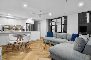 A modern kitchen with white cabinets, a dining area with a glass table, and a grey sofa with blue cushions. - Luxe 2 Bed Apt, Seconds to Beach & Minutes to City (Melbourne)