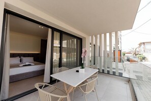 A balcony with a table and chairs, a bed, and a sliding glass door leading to a bedroom. - Aetheria North (Amaliada)