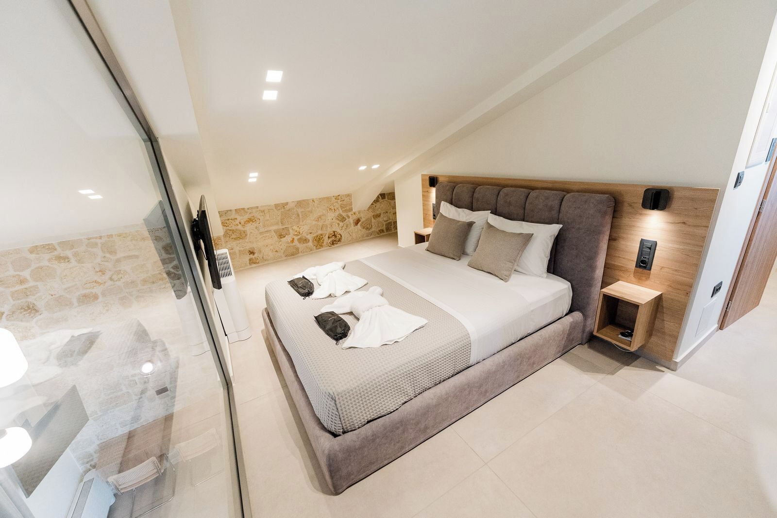 A modern bedroom with a stone accent wall, a large bed with a wooden headboard, and a bedside table with a lamp.
