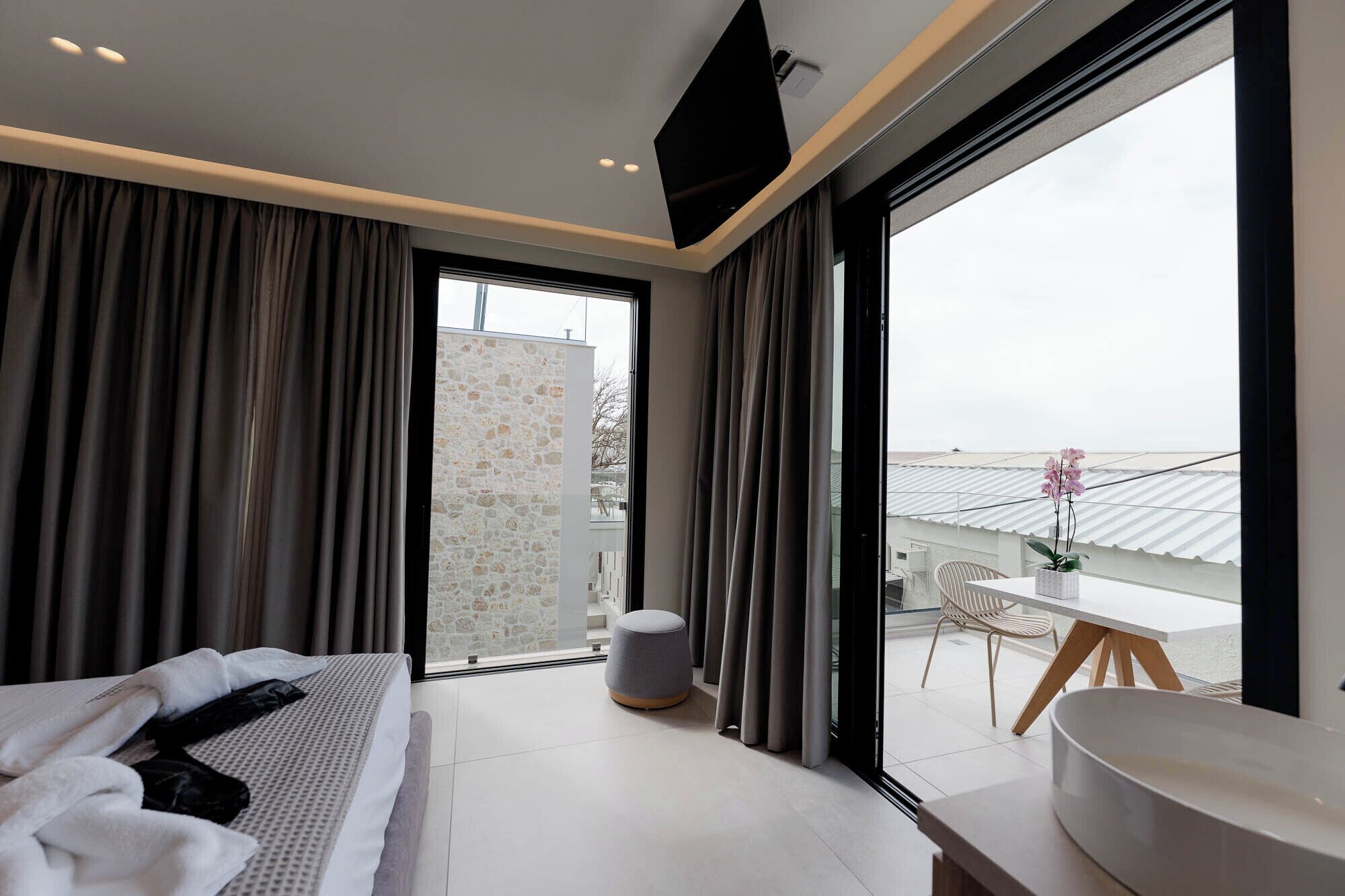 A modern hotel room with a bed, a small seating area, a balcony with a view, and a large window.