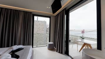 A modern hotel room with a bed, a small seating area, a balcony with a view, and a large window.