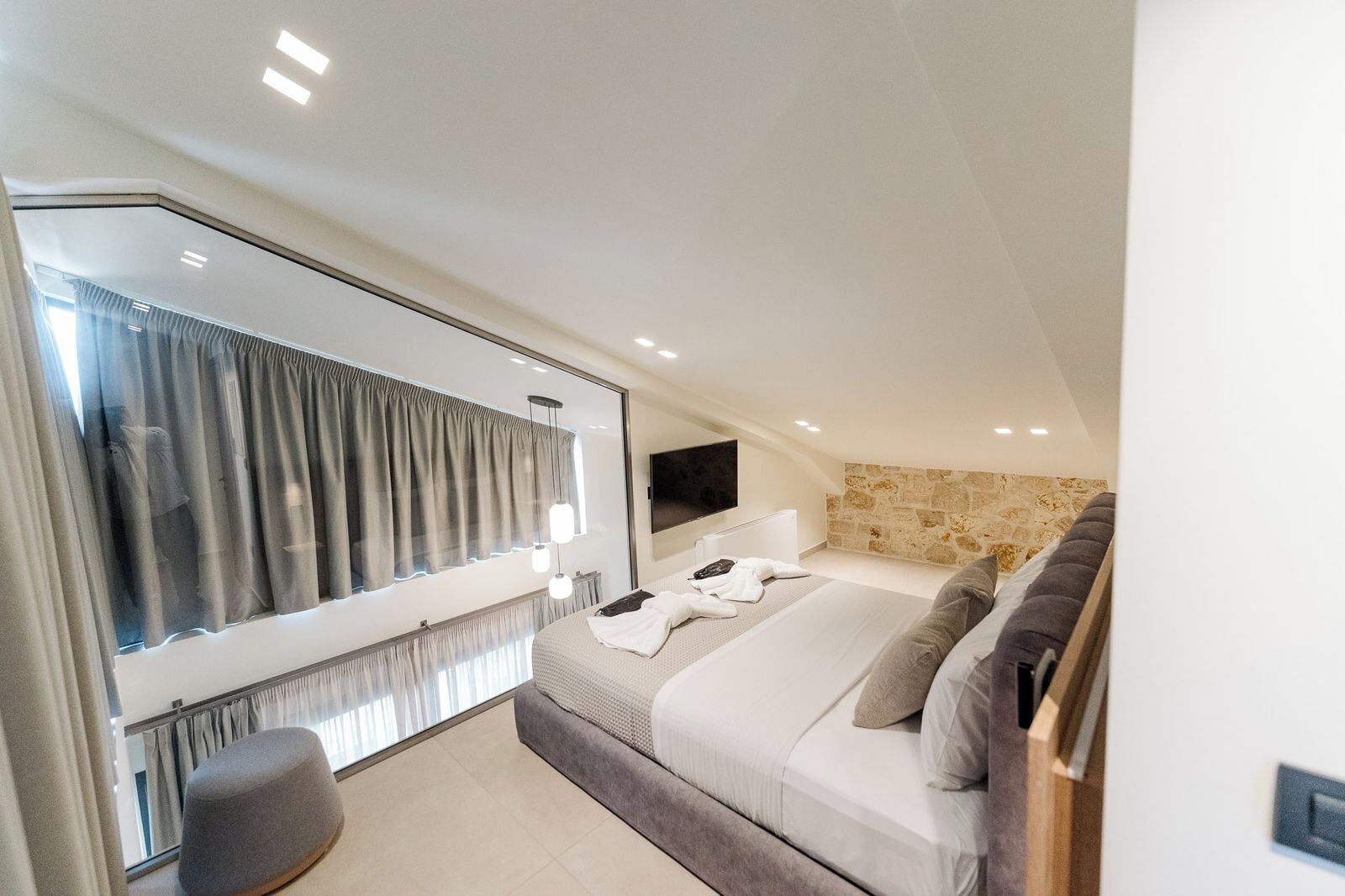 A modern bedroom with a large bed, a seating area, a television, and a stone wall.