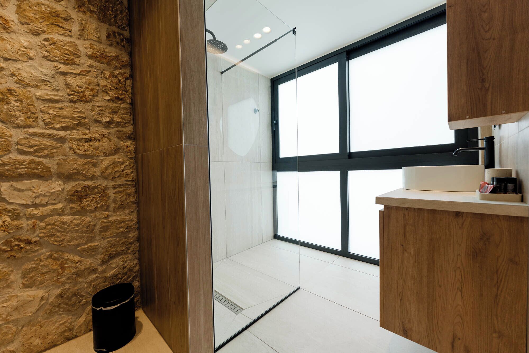 Un baño moderno con pared de piedra, cabina de ducha de vidrio y tocador de madera.