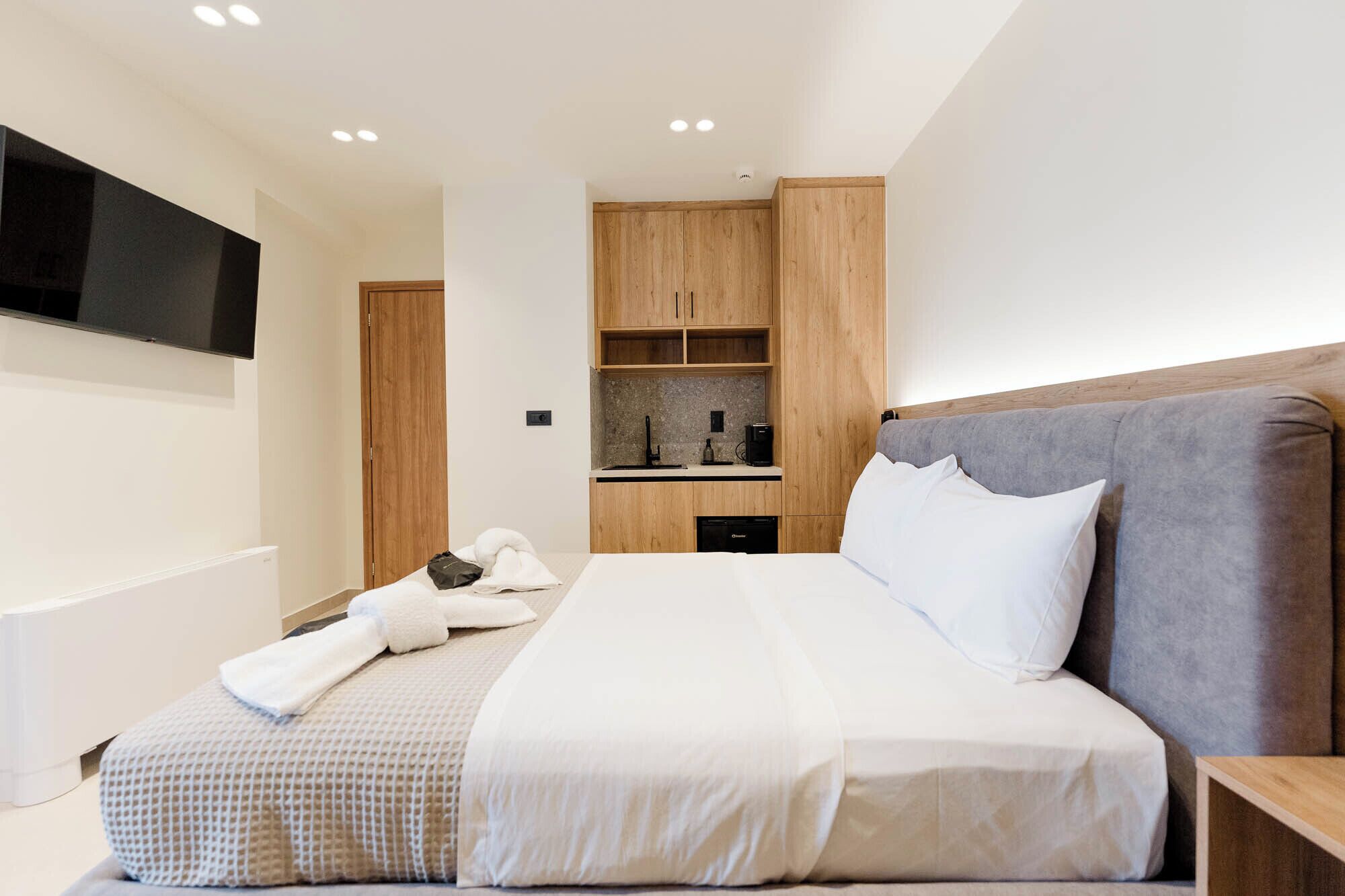 A modern hotel room with a large bed, a flat-screen TV mounted on the wall, a built-in kitchen area, and wooden cabinetry.