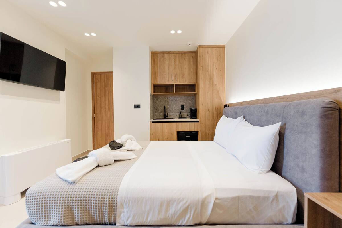 A modern hotel room with a large bed, a flat-screen TV mounted on the wall, a built-in kitchen area, and wooden cabinetry.