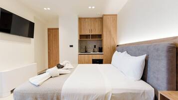 A modern hotel room with a large bed, a flat-screen TV mounted on the wall, a built-in kitchen area, and wooden cabinetry.