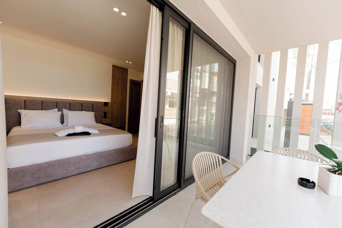 A modern hotel room with a bed, a sliding glass door to a balcony, a small table, and a chair.