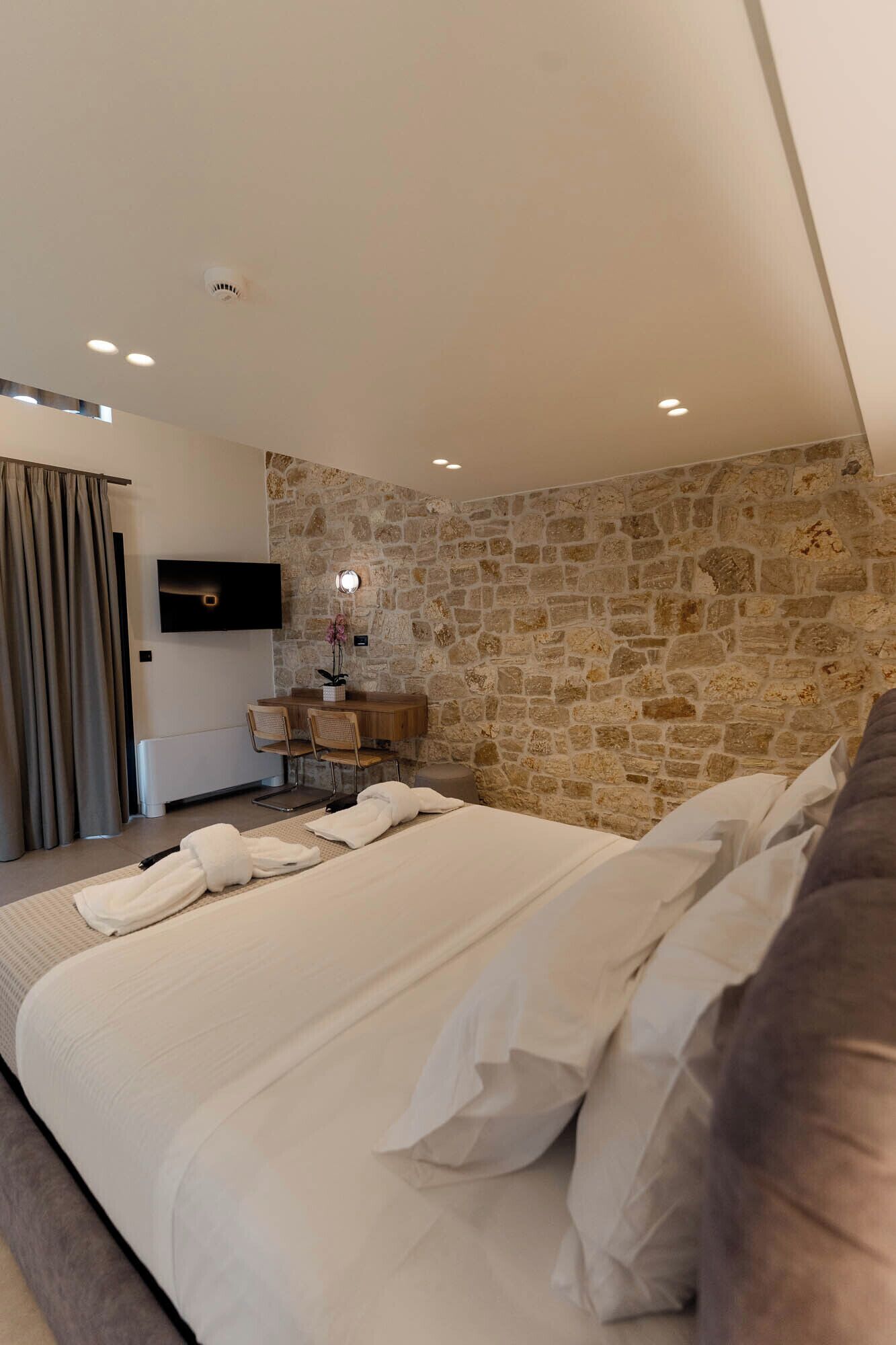 Een slaapkamer met een stenen muur, een flatscreen televisie, een bureau met stoel, en een bed met witte lakens.