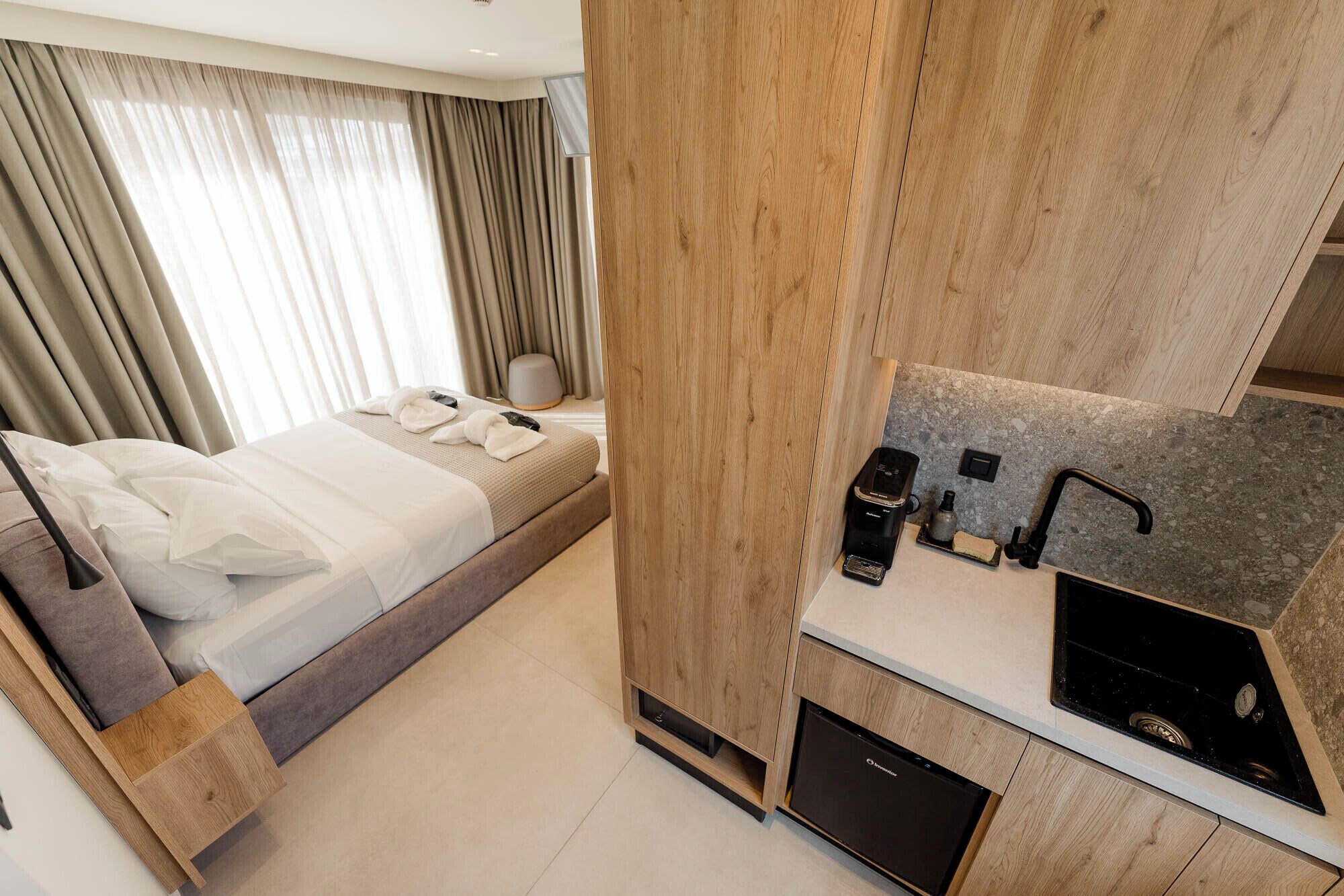 Habitación de hotel con cama, separador de madera, kitchenette con pileta y estufa, y ventana con cortinas.