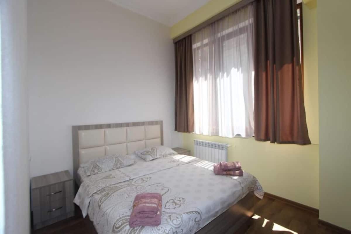 A bedroom with a bed, a nightstand, a window with curtains, and a radiator.
