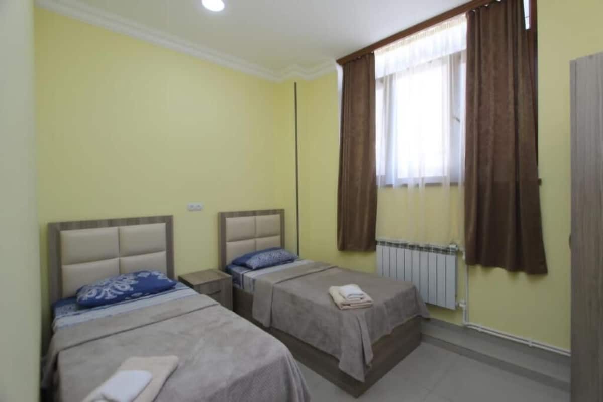 A room with two beds, a window with curtains, a radiator, and a door.