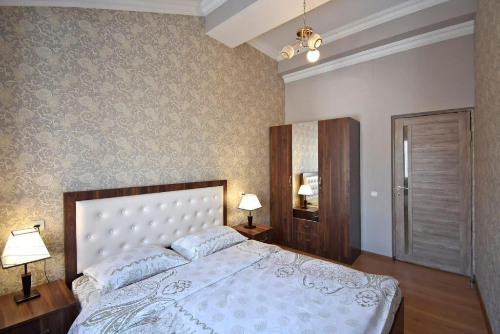 A bedroom with a large bed, a nightstand with a lamp, a wardrobe, and a door.