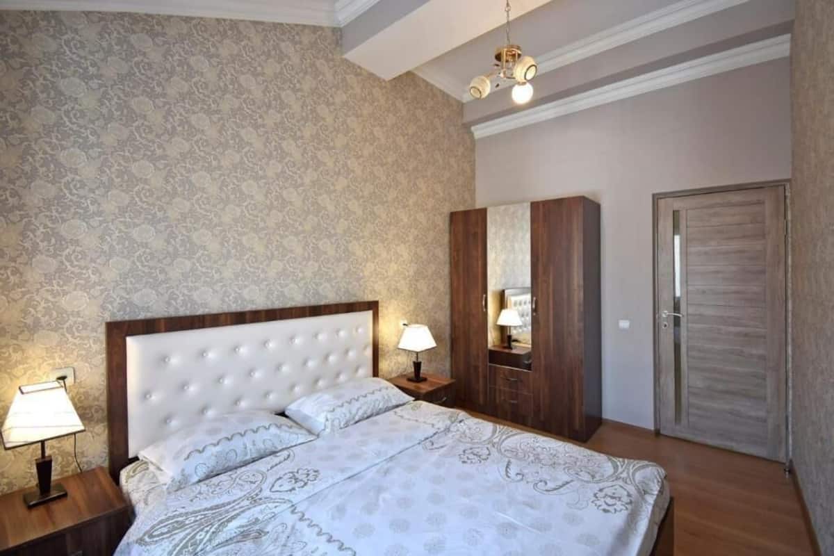 A bedroom with a large bed, a nightstand with a lamp, a wardrobe, and a door.