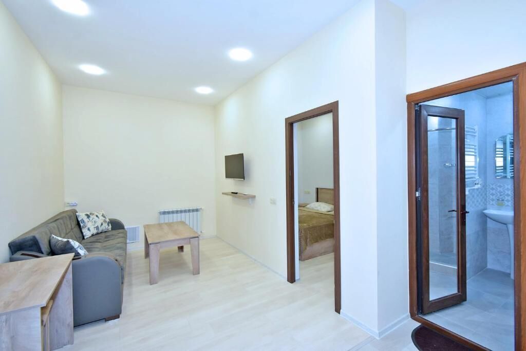 A modern apartment with a living area, a small table, a sofa, a TV mounted on the wall, and a bathroom visible through a glass door.