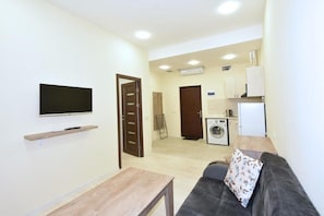 A living room with a sofa, a TV mounted on the wall, a wooden table, and a kitchen area with a washing machine.