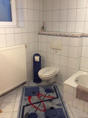 A bathroom with a toilet, a radiator, a bathtub, and a blue rug with abstract patterns. - Pure nature - relaxation in a quiet vacation apartment in the middle of the Harz Nature Park (Clausthal-Zellerfeld)