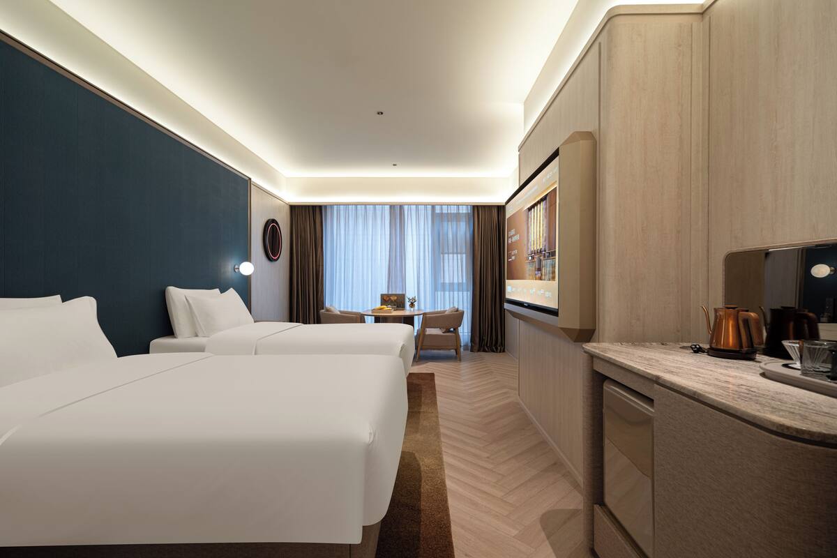 A hotel room with two beds, a desk, a chair, a kitchen area, and a large window with curtains.