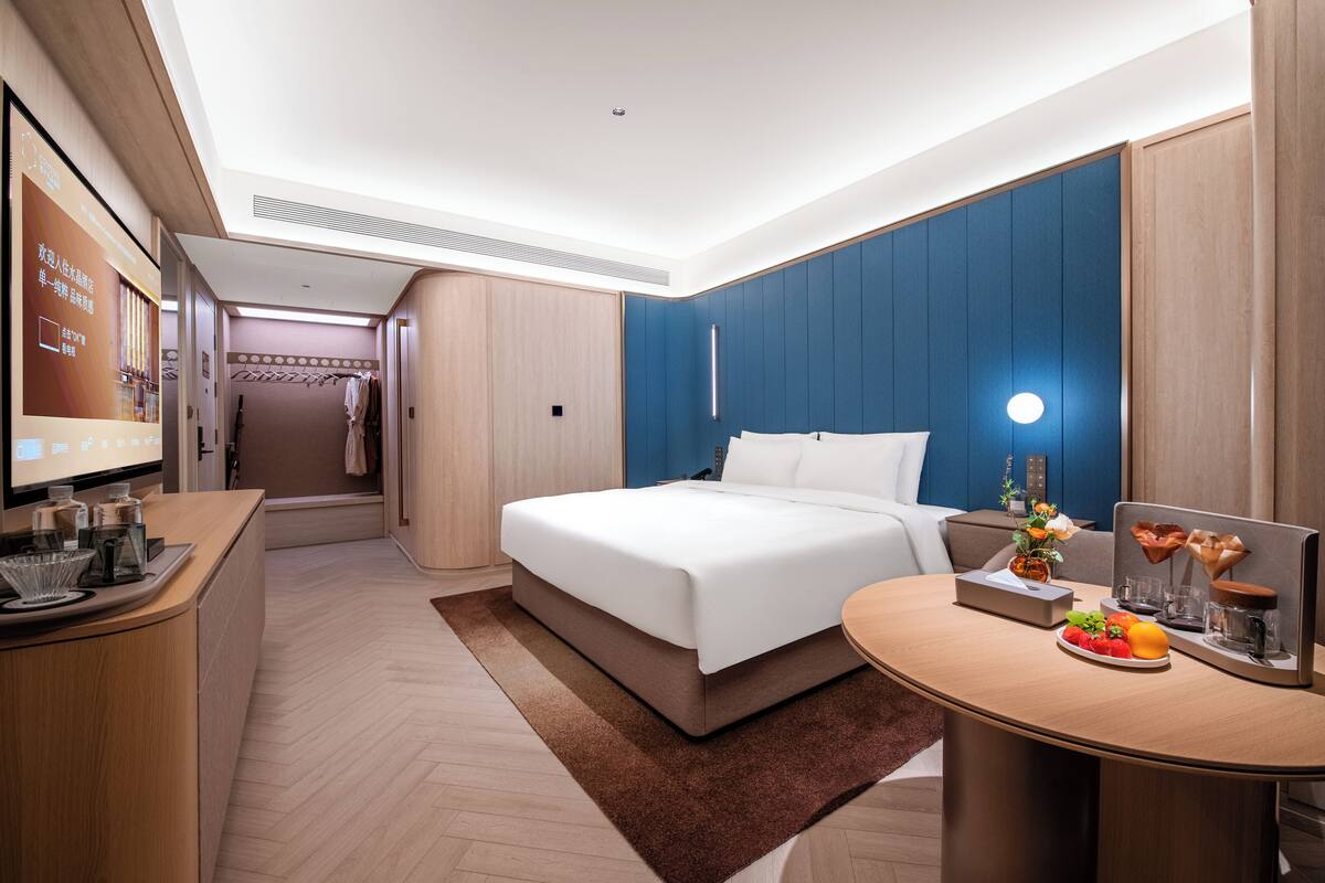 A hotel room with a bed, bedside table, and a small desk area with a fruit bowl.