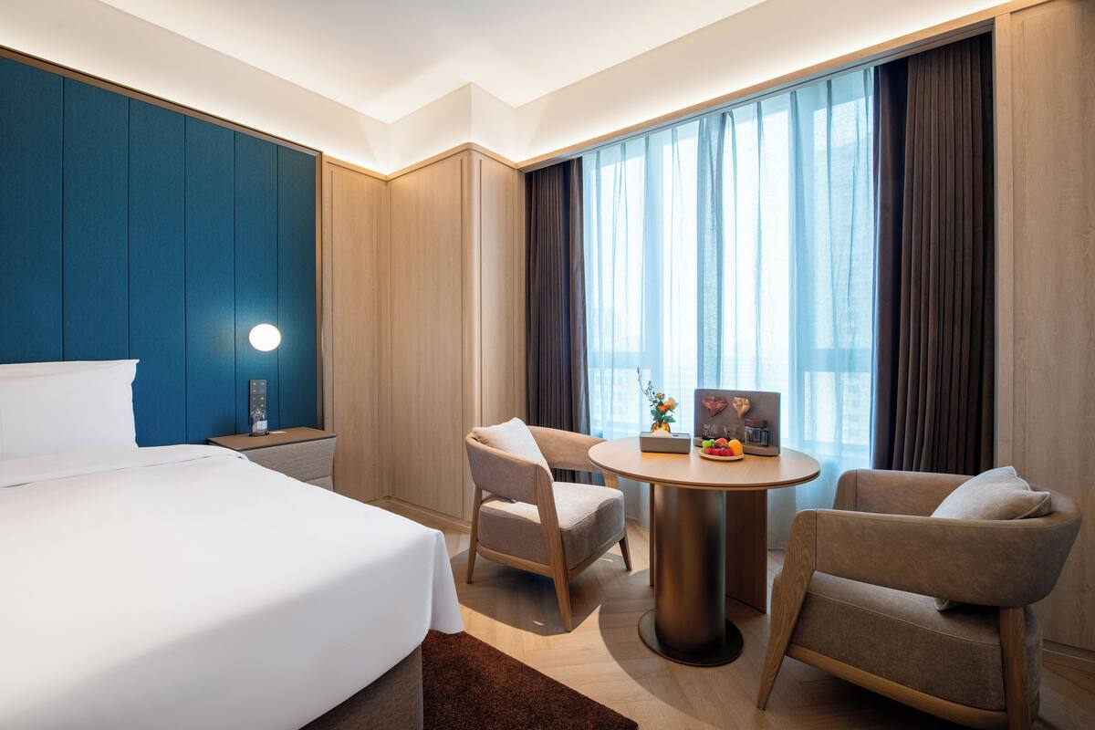 A hotel room with a bed, two armchairs, a small table, a nightstand, and a large window with curtains.