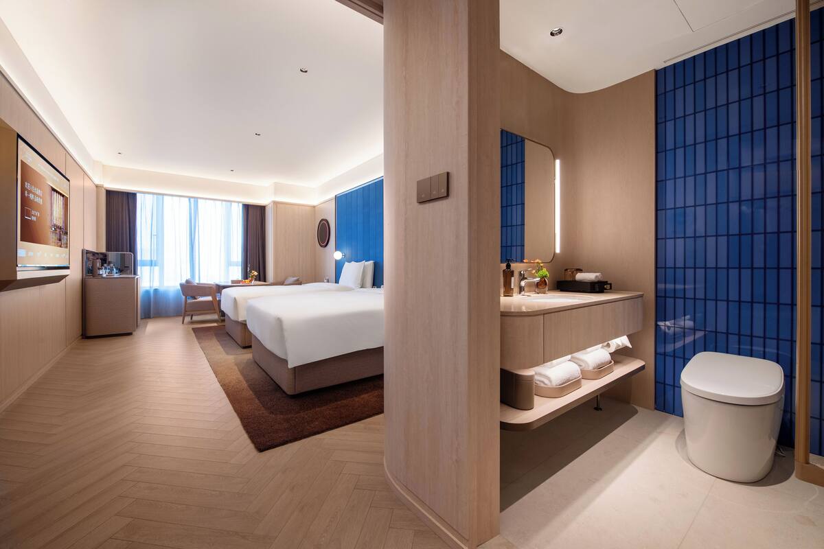 A hotel room with a bed, bedside table, a small desk with a chair, a bathroom with a toilet, and a large window.