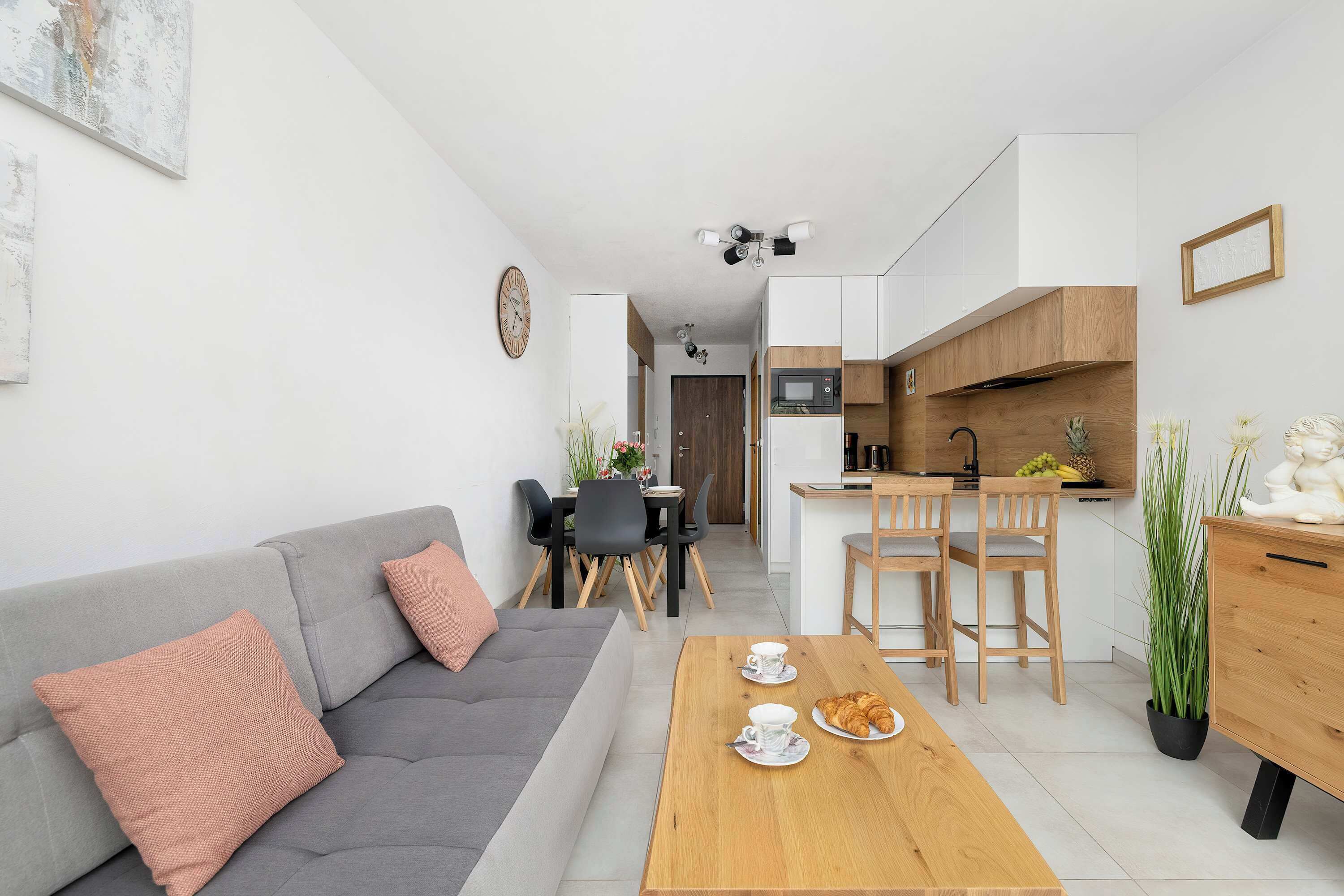 A modern apartment with a grey sofa, wooden dining table, kitchen area, and a view of the street.