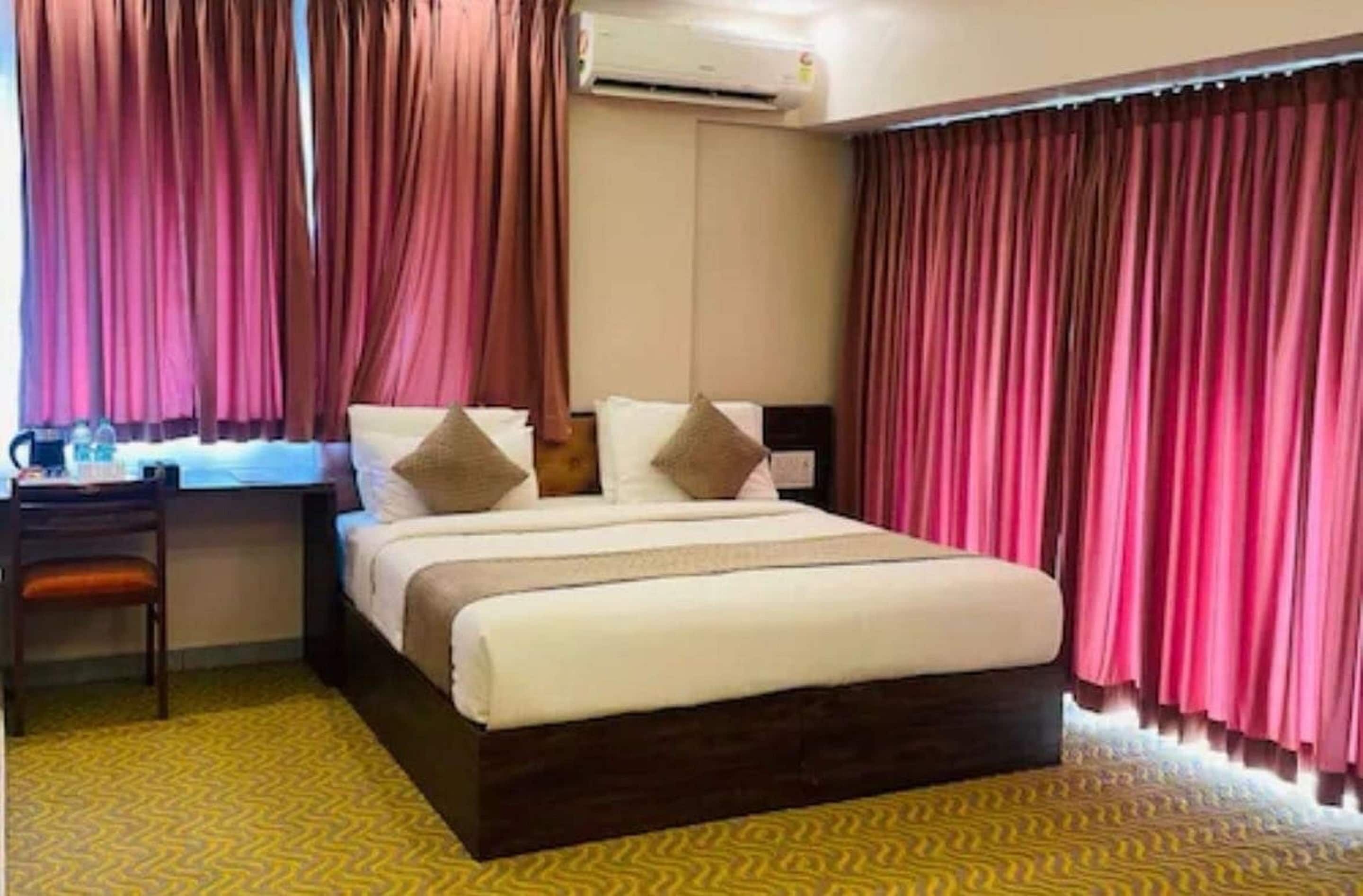 A hotel room with a bed, a desk with a chair, an air conditioner, and curtains.