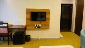 Quarto de hotel com cama, escrivaninha, televisão na parede e lixeira.