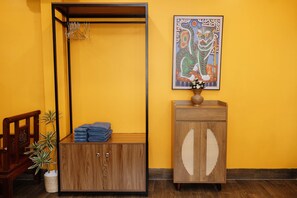 Ein Zimmer mit einem Holzkabinett, einer Kleiderstange, einem Stuhl, einer Topfpflanze und einem gerahmten Kunstwerk an der Wand.