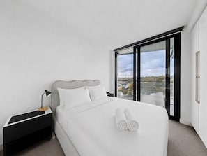 A modern hotel room with a large bed, a nightstand, a desk, and a view of the city through large windows. - Modern Living in the Heart of Hawthorn East (Melbourne)