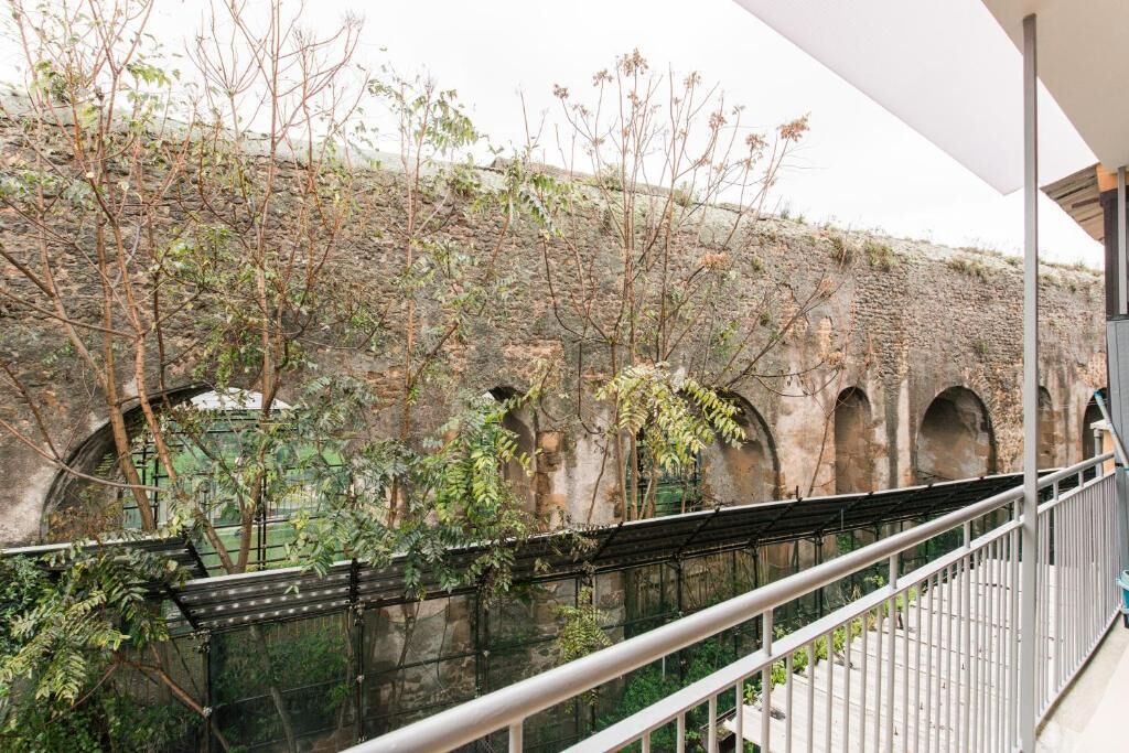 A balcony with a view of a stone wall and arches, surrounded by greenery.