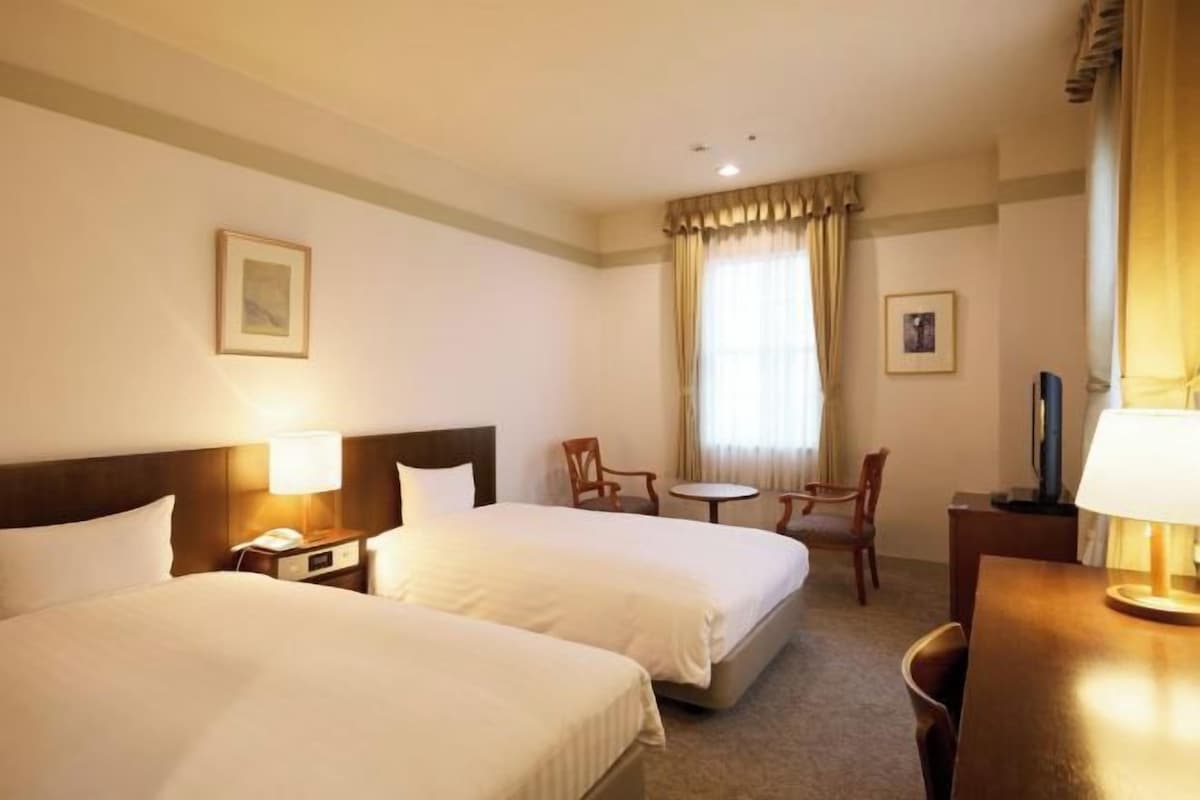 A hotel room with two beds, a small table, two chairs, a desk, and a window with curtains.