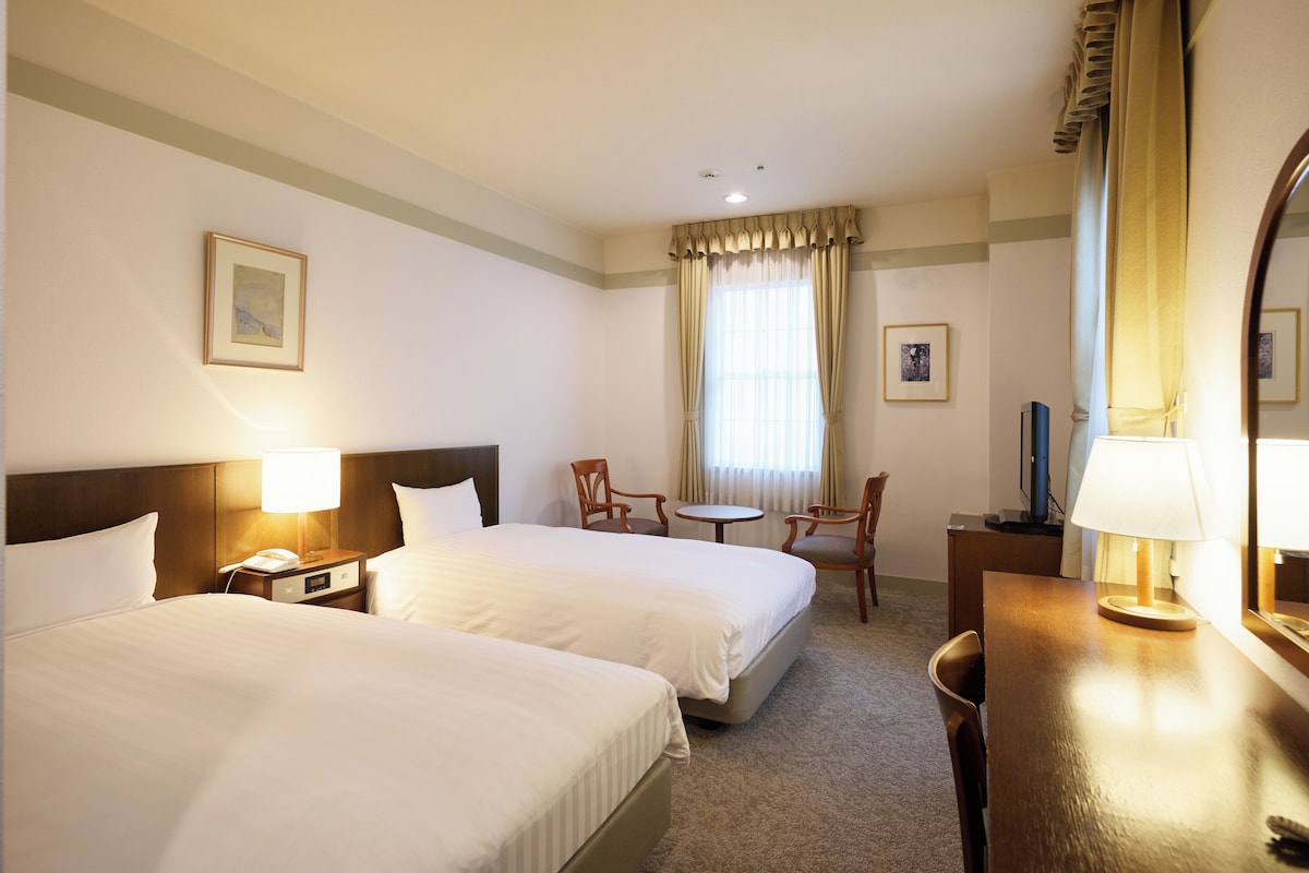 Ett hotellrum med två sängar, ett skrivbord, en stol, ett litet bord och en spegel.