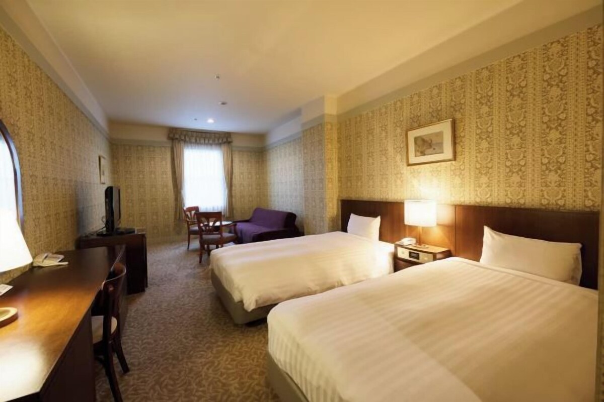 Quarto de hotel com duas camas, uma escrivaninha, uma cadeira, uma televisão e janela com cortinas.