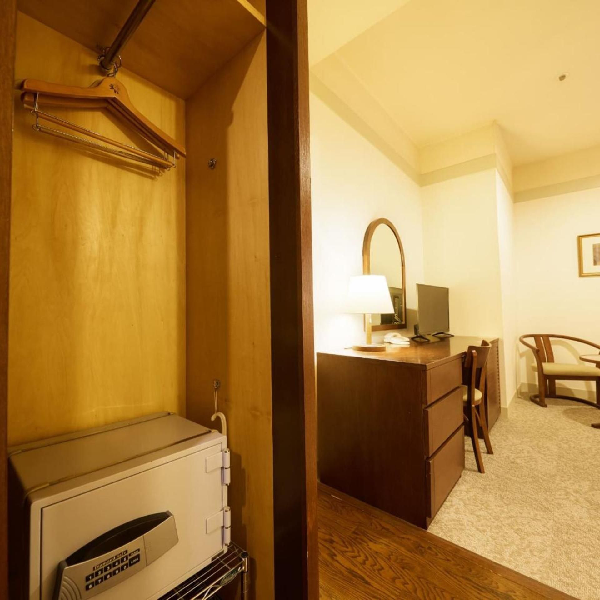 A hotel room with a wooden wardrobe, a desk with a lamp, a chair, and a small table.