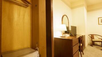 A hotel room with a wooden wardrobe, a desk with a lamp, a chair, and a small table.