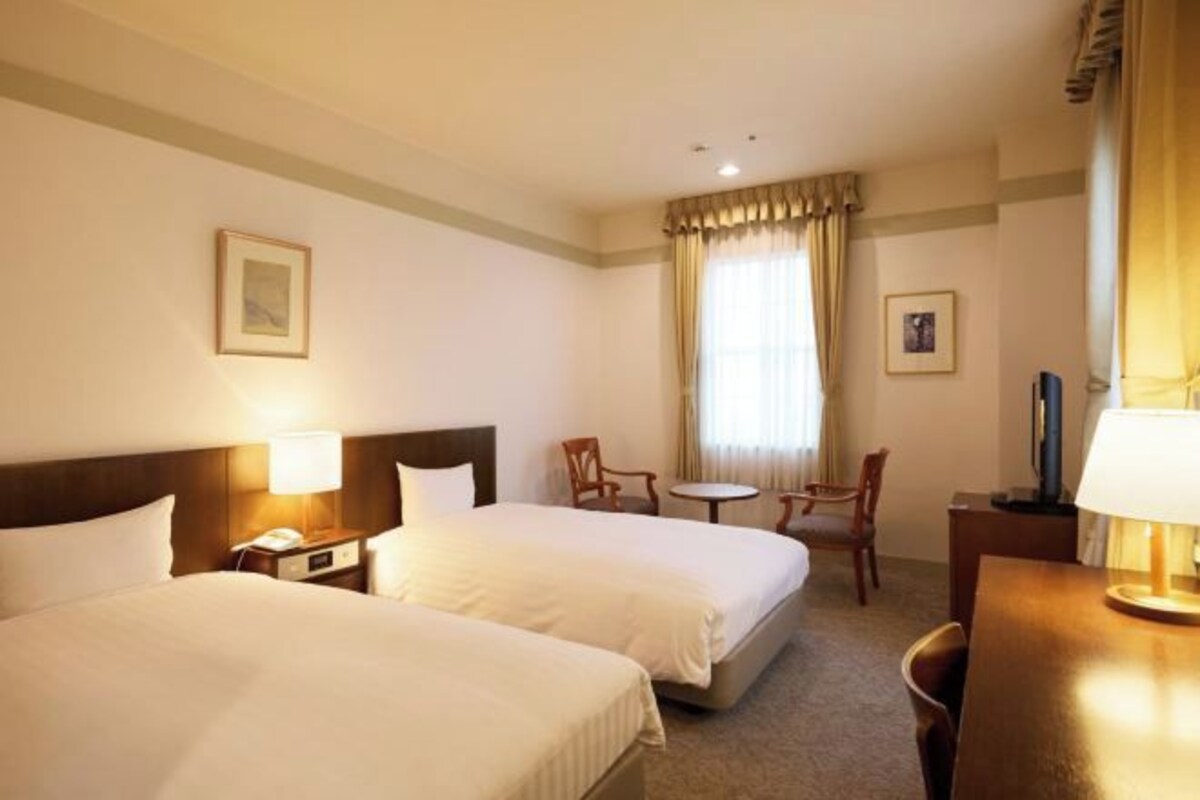 A hotel room with two beds, a small table, two chairs, a desk, and a window with curtains.
