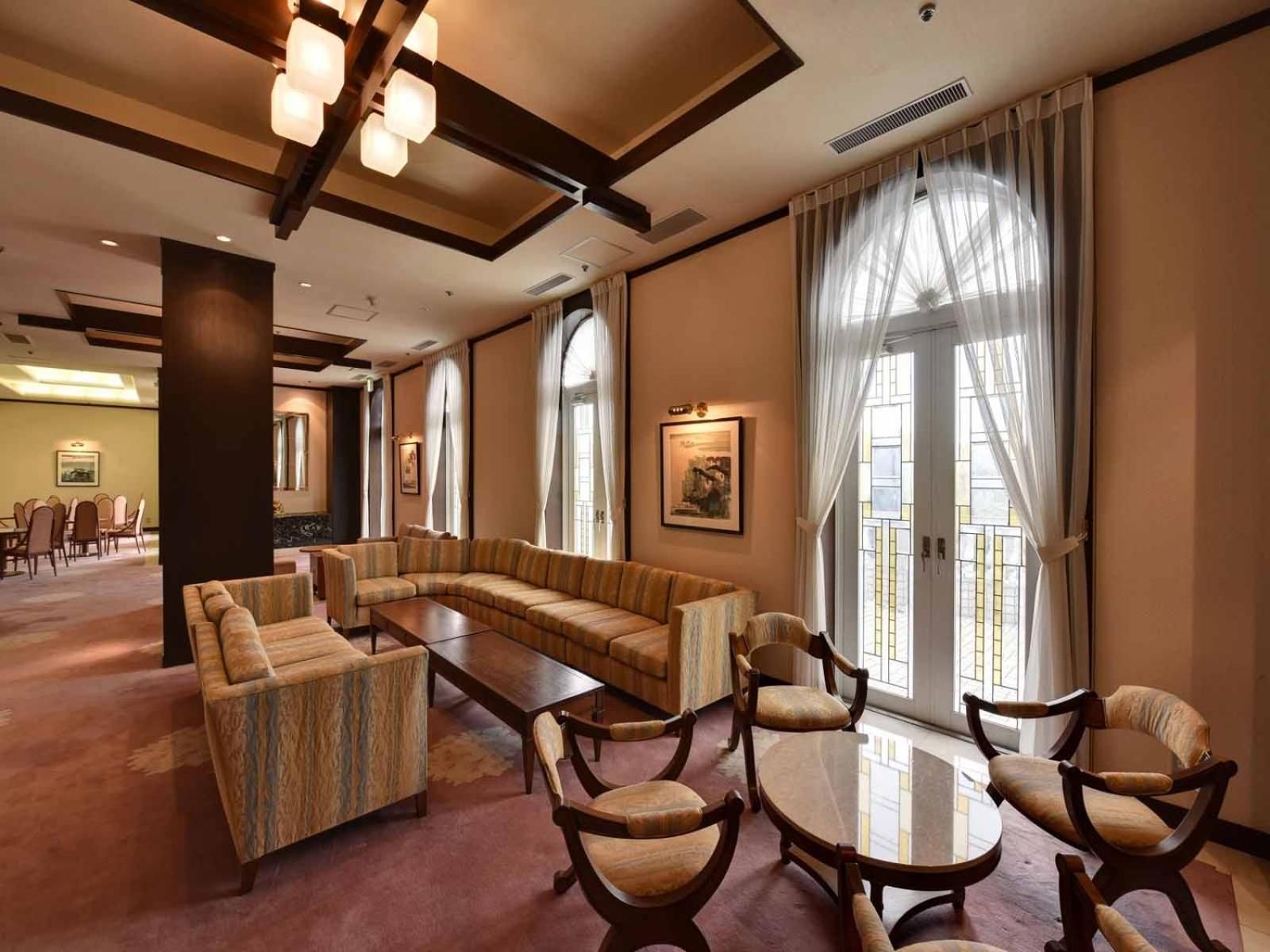 A hotel lobby with a sectional sofa, round table, and chairs, large windows with curtains, and a dining area in the background.
