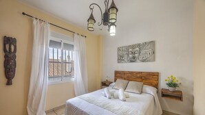 A bedroom with a bed, a window with curtains, a headboard, a wall-mounted artwork, a nightstand, and a small shelf with flowers. - Charming House in Vejer de la Frontera, Cádiz (Vejer de la Frontera)